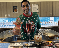 The founder wearing a green festive sweater smiling and holding up two colourful holiday cocktails behind an ethical mobile event bar displaying climate saving branding