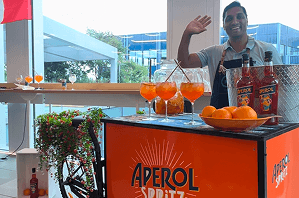 A smiling bartender waving from behind an Aperol Spritz branded drinks tricycle covered in bright orange cocktails and flowers