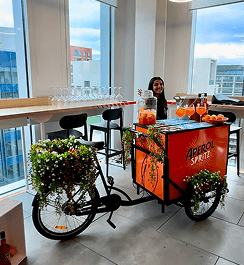 A smiling bartender waving from behind an orange Aperol Spritz branded drinks tricycle decorated with flowers in a bright modern office