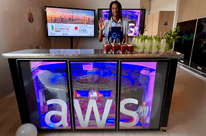 A bartender waving from behind a digital mobile event bar displaying the AWS logo across three video screens