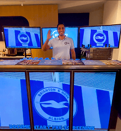 A bartender waving behind a digital mobile bar displaying the Brighton and Hove Albion football club logo on bright blue screens