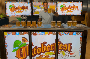 A smiling bartender holding up a glass of beer behind a digital event bar displaying Oktoberfest branding