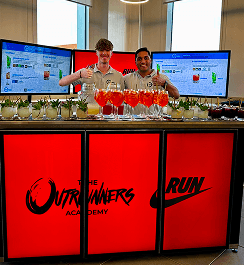 Two smiling bartenders giving thumbs up behind a digital pop up bar displaying Nike branding and lined with orange cocktails
