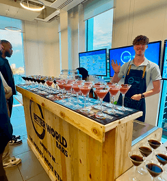 A smiling bartender waving from behind a reclaimed wood pop up bar lined with pink cocktails