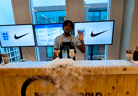 A bartender waving behind a wooden mobile bar covered in dry ice smoke with two digital screens displaying a brand logo