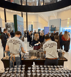 Two bartenders wearing shirts that read aiming to be the worlds most ethical bar serving drinks to a large crowd in a modern office atrium