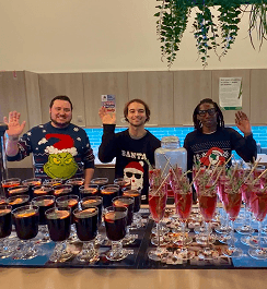 Three bartenders wearing festive Christmas sweaters waving from behind an office pop up bar covered in glasses of red holiday cocktails