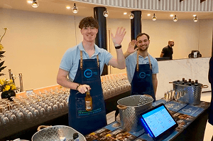 Two smiling bartenders waving from behind a pop up event bar in a bright modern corporate reception area