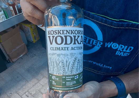 A bartender wearing a denim apron holding a bottle of climate action vodka at a sustainable festival bar