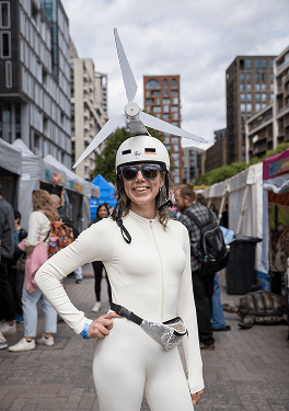 A smiling woman at an outdoor festival wearing a white outfit and a helmet with a wind turbine attached to the top