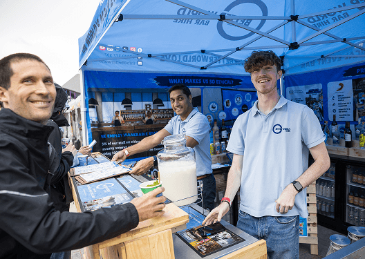 Two bartenders serving drinks to festival attendees from a wooden mobile bar under a blue canopy