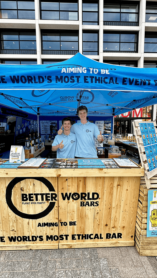 Three smiling bartenders standing behind a wooden event bar serving drinks to festival attendees