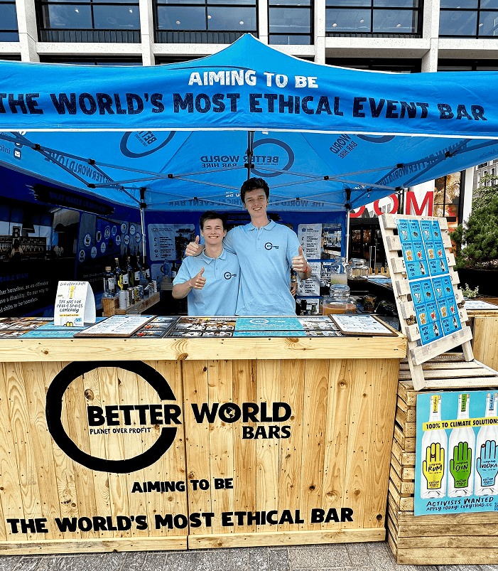 Two smiling bartenders giving thumbs up behind a wooden pop up event bar under a blue branded canopy at an outdoor sustainability festival if