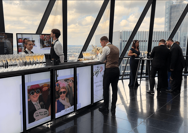 A bartender opening a bottle of champagne behind a digital event bar while formally dressed wedding guests mingle near the glass windows of the Gherkin