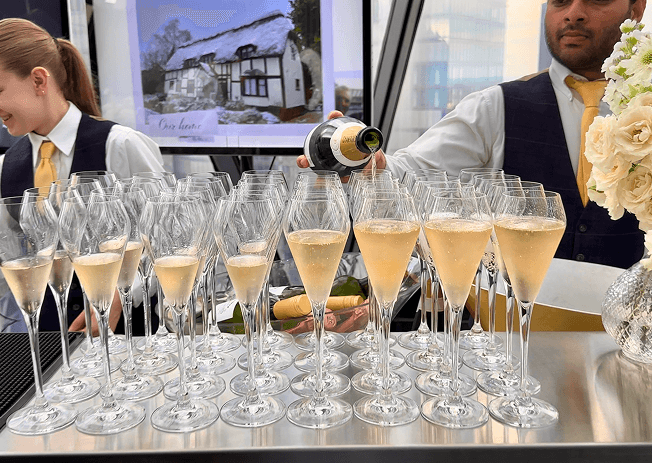 A bartender pouring champagne into a large group of flute glasses on a mobile bar with a digital screen displaying a photo of a house in the background