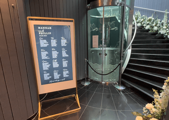 A digital wedding seating plan sign for Hannah and Phillip standing next to a glass elevator and a black staircase decorated with white flowers