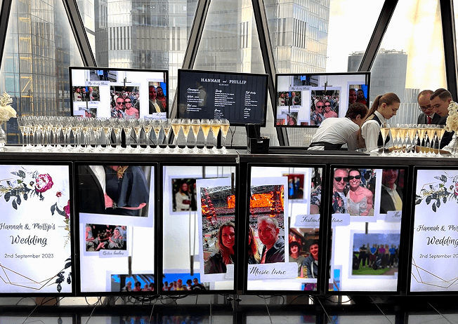 Three venue staff members preparing drinks behind a premium digital mobile bar displaying personalized wedding photos with the London skyline in the background