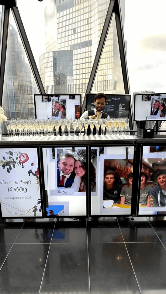 A bartender pouring champagne into multiple flute glasses lined up on a digital wedding bar displaying photos of the bride and groom at the Gherkin building