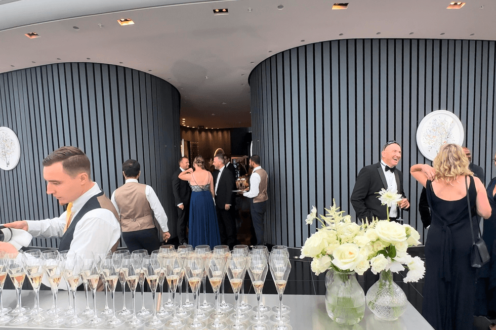 A wide view of wedding guests in suits and evening dresses mingling and drinking champagne inside the top floor of the Gherkin building