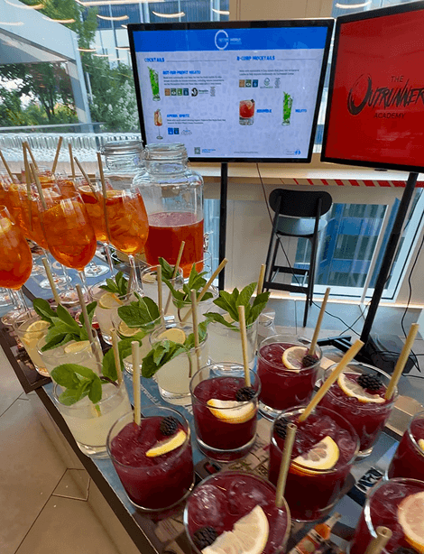A close up of multiple fresh cocktails and mocktails including Aperol Spritz and Mojitos lined up on a mobile event bar with digital menu screens in the background