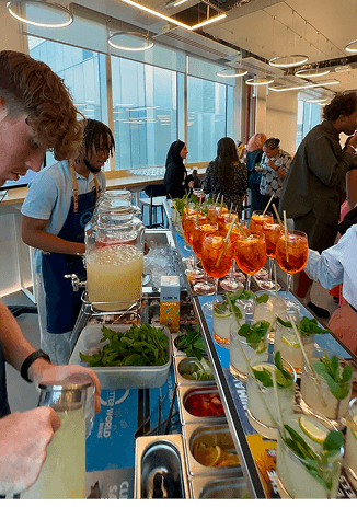 Two bartenders preparing fresh drinks behind a mobile bar equipped with glass beverage dispensers while guests mingle in the background