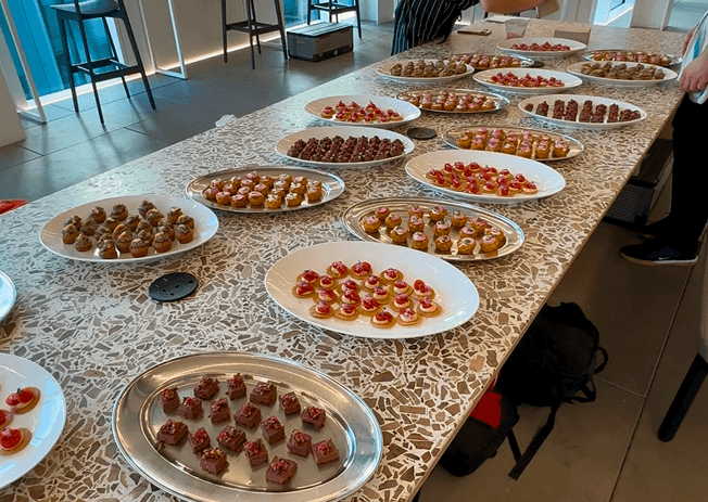 Fresh cocktails lined up on an event bar with fresh fruit garnishes alongside a long table filled with plates of catered food