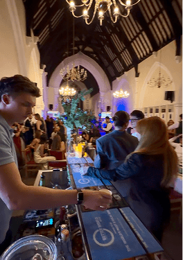 A bartender serving drinks to guests wearing formal attire at a brightly lit mobile event bar inside a large hall