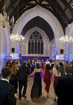 A large group of guests holding hands and participating in a traditional Scottish dance on a wooden floor at a charity ball