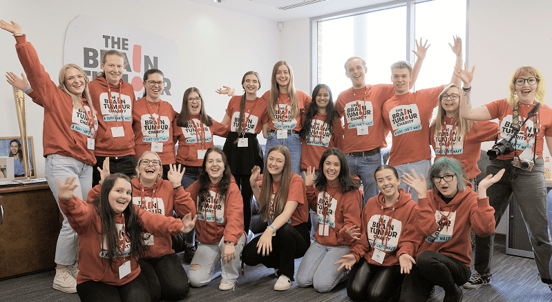 A large group of smiling people wearing red The Brain Tumour Charity hoodies waving at the camera representing the incredible charitable causes supported by our sustainable event drinks