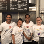 Four smiling staff members wearing white charity t shirts standing behind a bar next to a charity cocktail sign representing our commitment to employing vulnerable staff