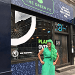 A smiling person wearing a bright green wig and a green dress standing outside The Green Vic pub representing the funds we raised for local charities