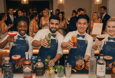 Four smiling professional bartenders raising their cocktail glasses behind a white marble bar representing our fully licensed and insured wedding bar team
