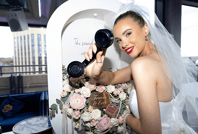 A smiling bride holding a vintage black telephone receiver next to a beautiful floral display representing the simple process of how to book your bespoke wedding bar