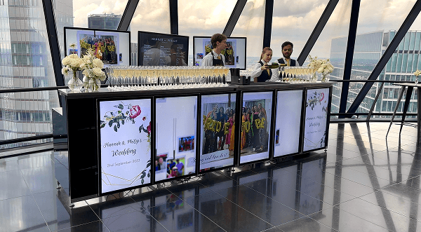 Three bartenders pouring champagne behind a premium luxury digital wedding bar displaying personalized photos of the couple representing our high tech showstopper for modern wedding receptions