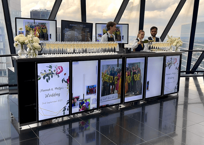 Three bartenders pouring champagne behind a glowing digital mobile wedding bar displaying personalised photos inside a modern glass building overlooking the city representing our bespoke quotes and unforgettable experiences for your special day