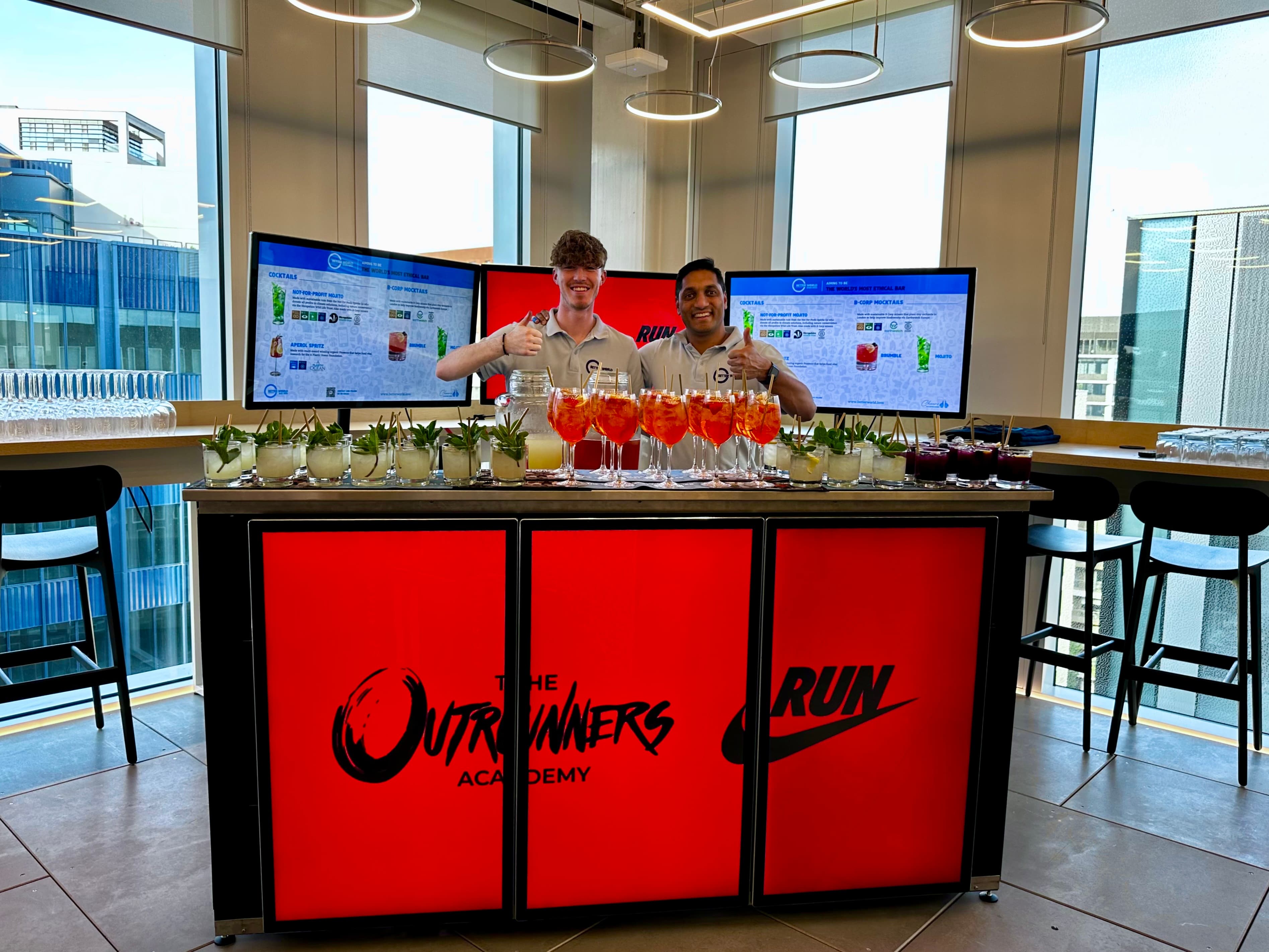 Two professional bartenders smiling behind a sustainable digital pop up event bar displaying Nike branding with freshly prepared cocktails ready for a corporate event.