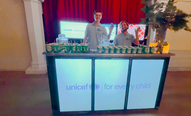 Two smiling team members standing behind a glowing digital mobile event bar displaying the UNICEF logo across its front panels representing our premium hospitality and mission driven corporate partnerships
