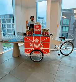A professional bartender waving behind a compact drinks tricycle displaying custom Pimms branding ready to serve welcome drinks at a product activation