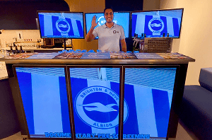 A professional bartender smiling and waving behind a high tech digital screen bar displaying custom Brighton and Hove Albion branding for a VIP documentary event