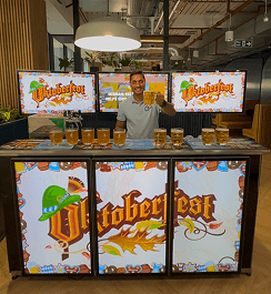 A professional bartender raising a glass of beer behind a digital event bar displaying immersive Oktoberfest branding for a corporate office party