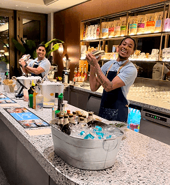Two expert mixologists shaking sustainable cocktails behind a transformed office space counter providing premium ethical hospitality for a staff event