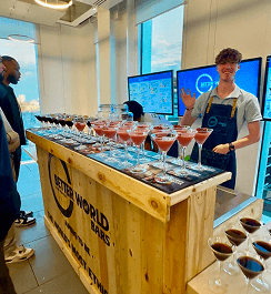 A professional bartender smiling and waving behind a reclaimed wood pop up bar filled with sustainable cocktails for a corporate office party