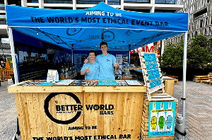 Two professional team members giving a thumbs up behind a handcrafted wooden mobile bar set up under a branded gazebo for an outdoor sustainability event
