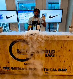 A professional bartender waving from behind a rustic wooden event bar featuring a dry ice display and digital screens for a corporate brand activation
