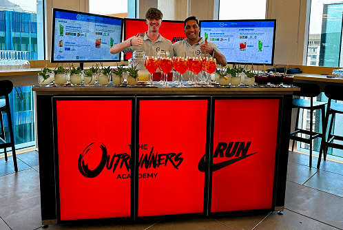 Two professional team members thumbs up behind a sustainable digital pop up bar displaying Nike branding with freshly made cocktails ready for service.