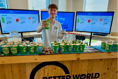 A professional bartender holding up an eco cup and smiling behind a rustic wooden pop up bar displaying our sustainable mission statement.