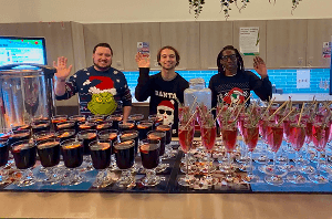 Three professional team members in festive jumpers smiling and waving behind a pop up event bar serving winter cocktails.