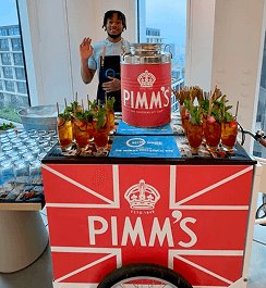 A professional bartender smiling and waving behind a branded drinks tricycle serving premium summer cocktails at a corporate event.