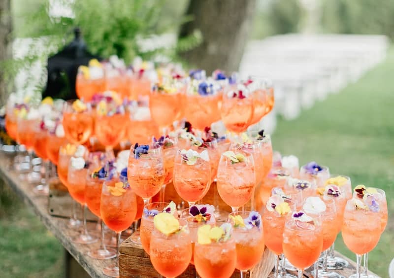 Dozens of orange spritzes garnished with edible flowers set out across a rustic wooden trestle bar at an outdoor wedding, fully installed and ready for guests, representing our seamless wedding bar delivery and setup